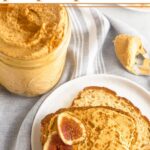 Paleo pumpkin pie butter spread on a piece of toast with a slice of fig on it. It is on a white plate next to a jar of pumpkin pie butter, and a spoon with pumpkin pie butter on it. Behind it is half an apple and a plate of sliced apples.