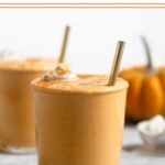 Pumpkin pie milkshake in a glass cup topped with whipped cream and cinnamon with a straw in it. Behind it is another cup along with a pumpkin and an ice cream scoop filled with ice cream.