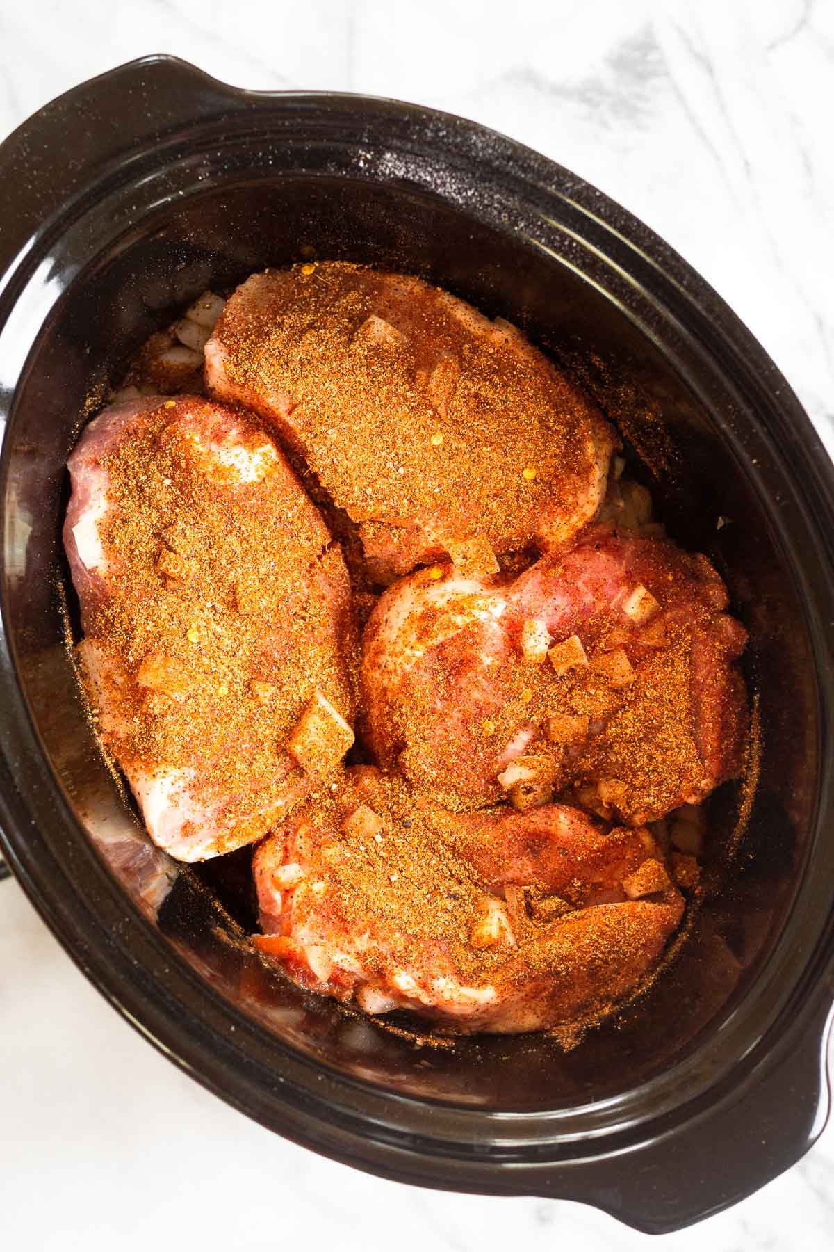 Crockpot filled with raw seasoned pork chops and a few diced onions.