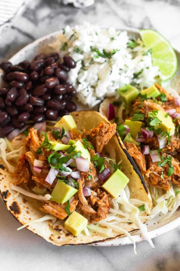 A plate of pulled pork tacos with cabbage, red onion, avocado, and cilantro with rice and beans on the side.