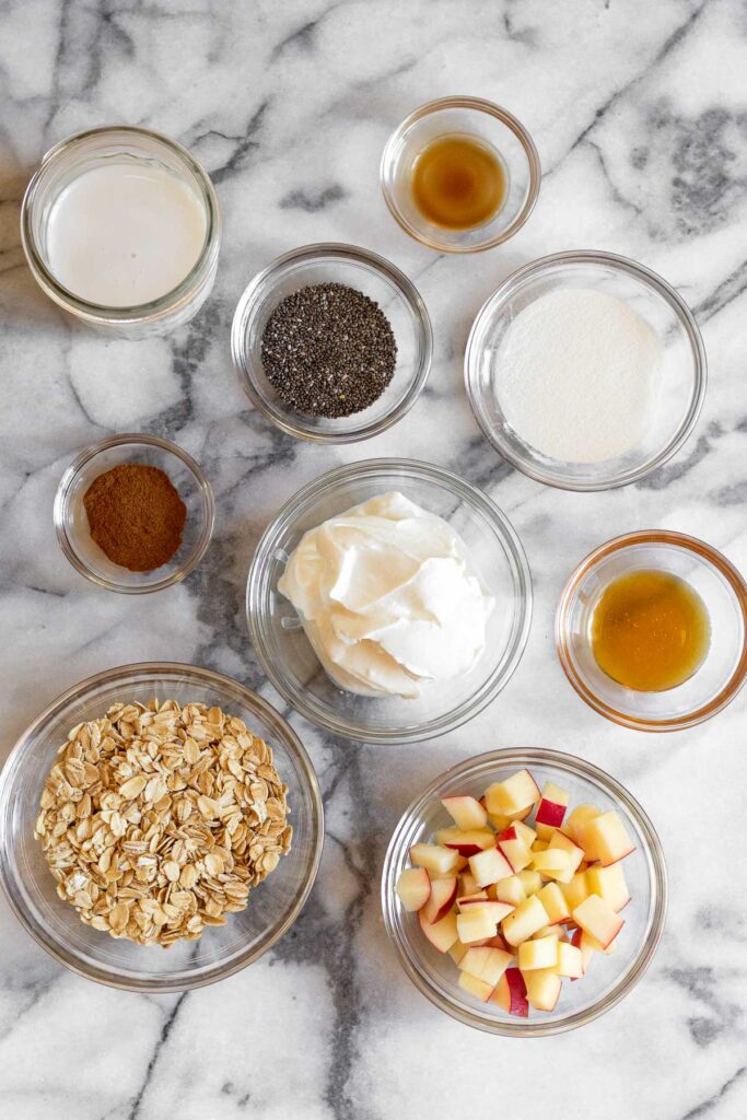 Overhead shot of a jar of almond milk, bowl of vanilla extract, bowl of collagen, bowl of honey, bowl of diced apples, bowl of oats, bowl of yogurt, bowl of cinnamon, and a bowl of chia seeds.