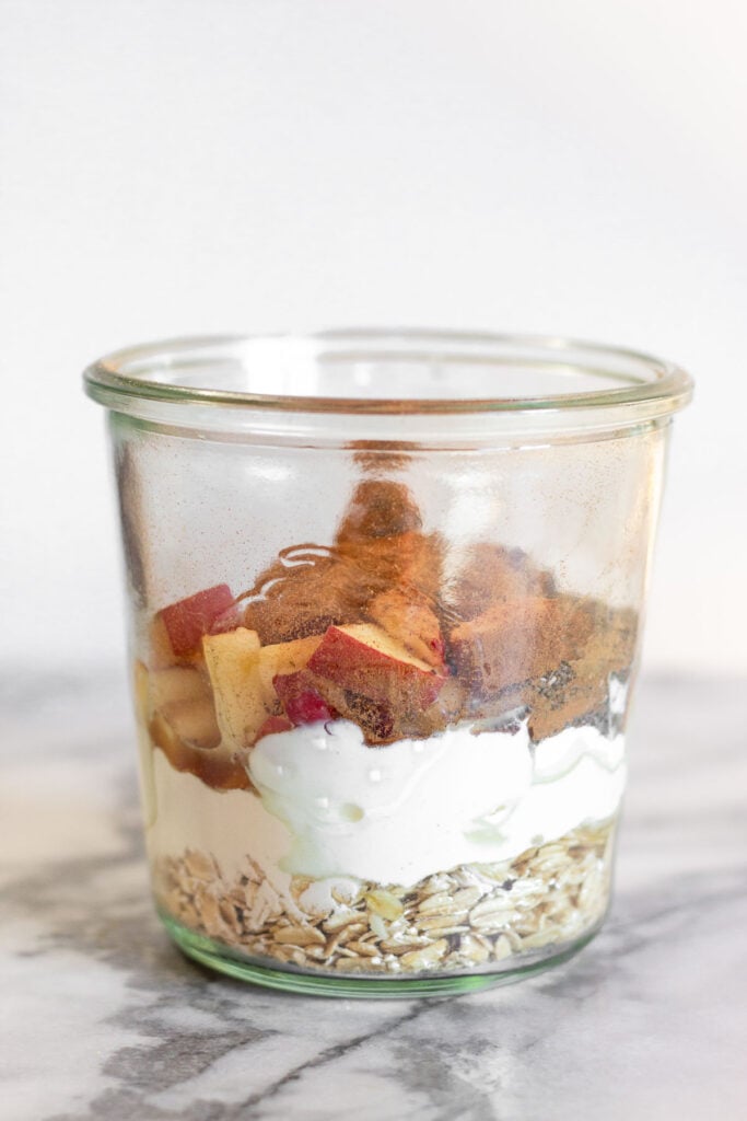 Jar filled with rolled oats, yogurt, diced apples, and cinnamon.