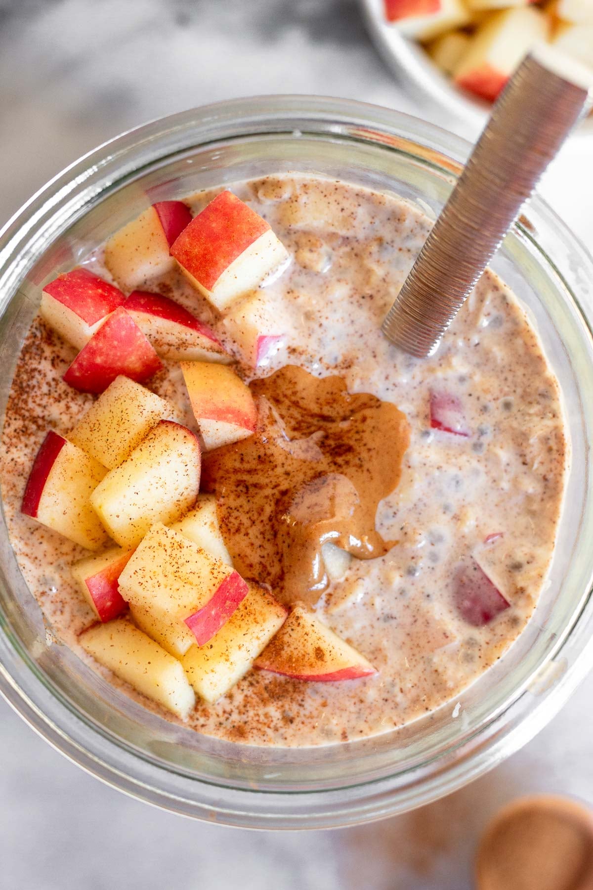 Apple cinnamon overnight oats in a jar topped with diced apples, peanut butter, and cinnamon and a spoon in the jar.