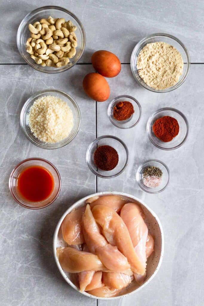 Overhead shot of a bowl of cashews, 2 eggs, bowl of coconut flour, a few bowls of spices, bowl of raw chicken tenderloins, bowl of hot sauce, and a bowl of shredded coconut.