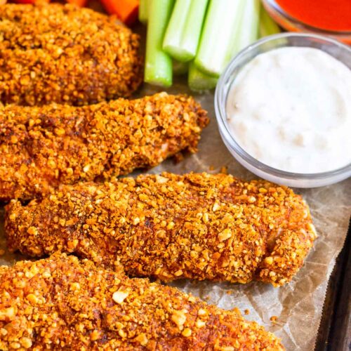 Buffalo chicken tenders on a baking sheet with carrots, celery, of bowl of buffalo sauce, and a bowl of blue cheese dressing.