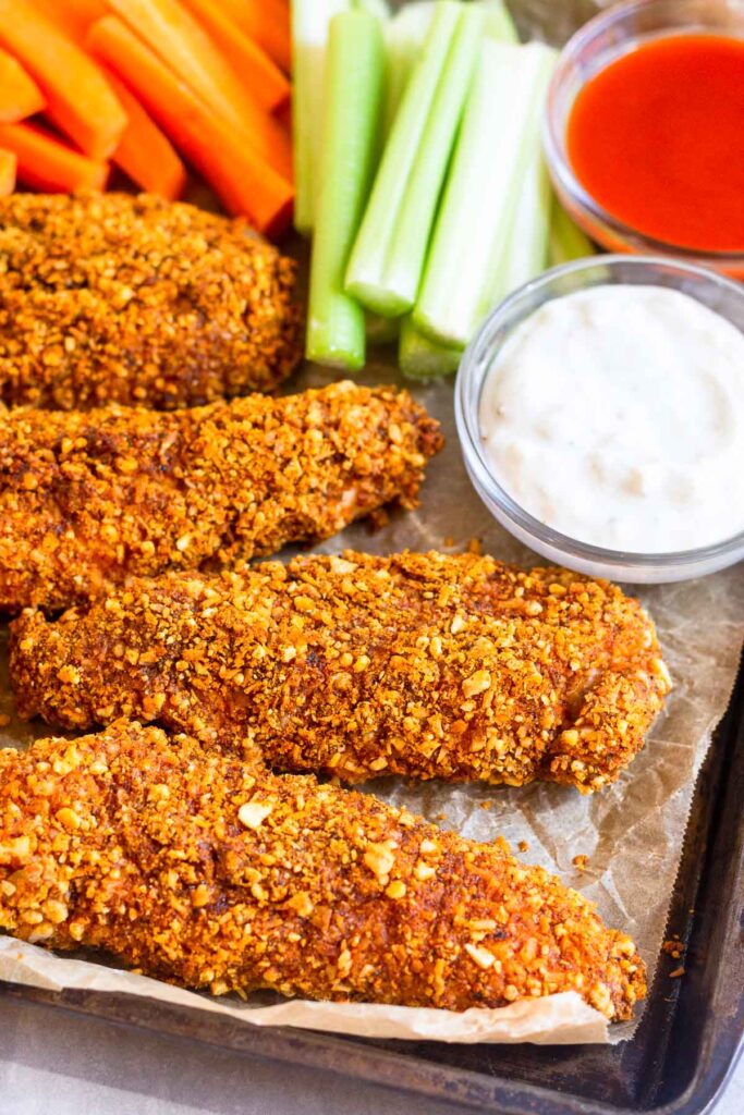 Buffalo chicken tenders on a baking sheet with carrots, celery, of bowl of buffalo sauce, and a bowl of blue cheese dressing.