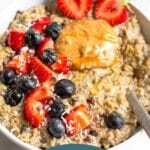 Bowl of cauliflower oats topped with strawberries, blueberries, peanut butter, and honey with a spoon coming out of the bowl. Behind it is a small ramekin of sliced strawberries.