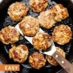 Chicken apple sausage patties in a large cast iron skillet. Two of them are lifted up on a spatula. The pan is sitting a brown linen with apples around it.
