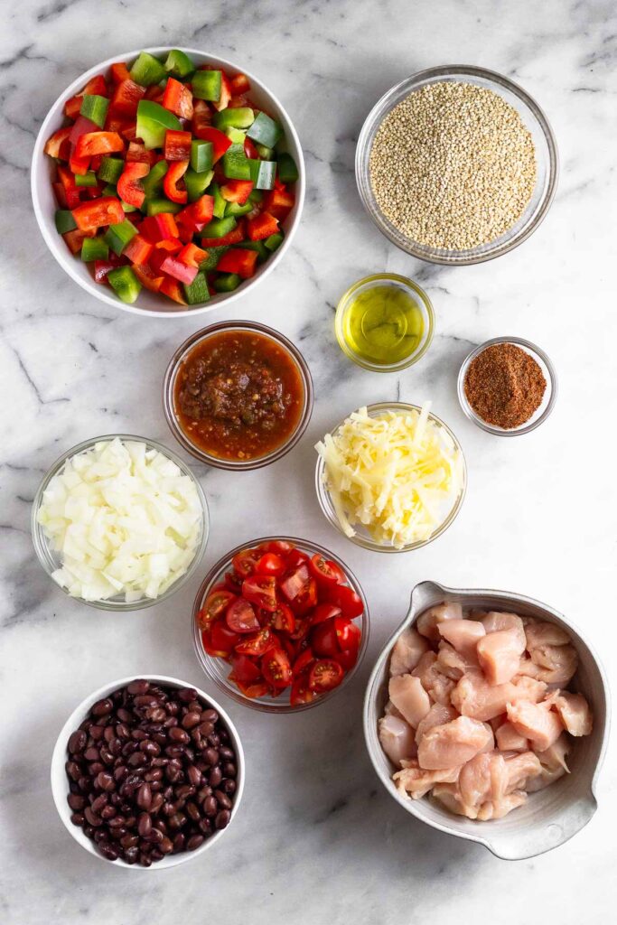 White counter with a bowl of diced peppers, bowl of dry quinoa, bowl of olive oil, bowl of spices, bowl of shredded cheese, bowl of diced raw chicken, bowl of black beans, bowl of diced tomatoes, bowl of diced onion, and a bowl of salsa.