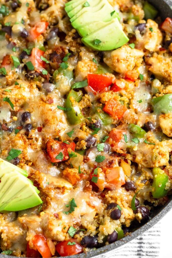 Close up of a skillet filled with chicken, quinoa, veggies, and melted cheese. It is topped with cilantro and avocado.