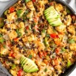 Chicken quinoa skillet in a large pan with veggies and topped with melted cheese topped with avocado. Around the pan is an half of avocado and a bowl of sliced lime.