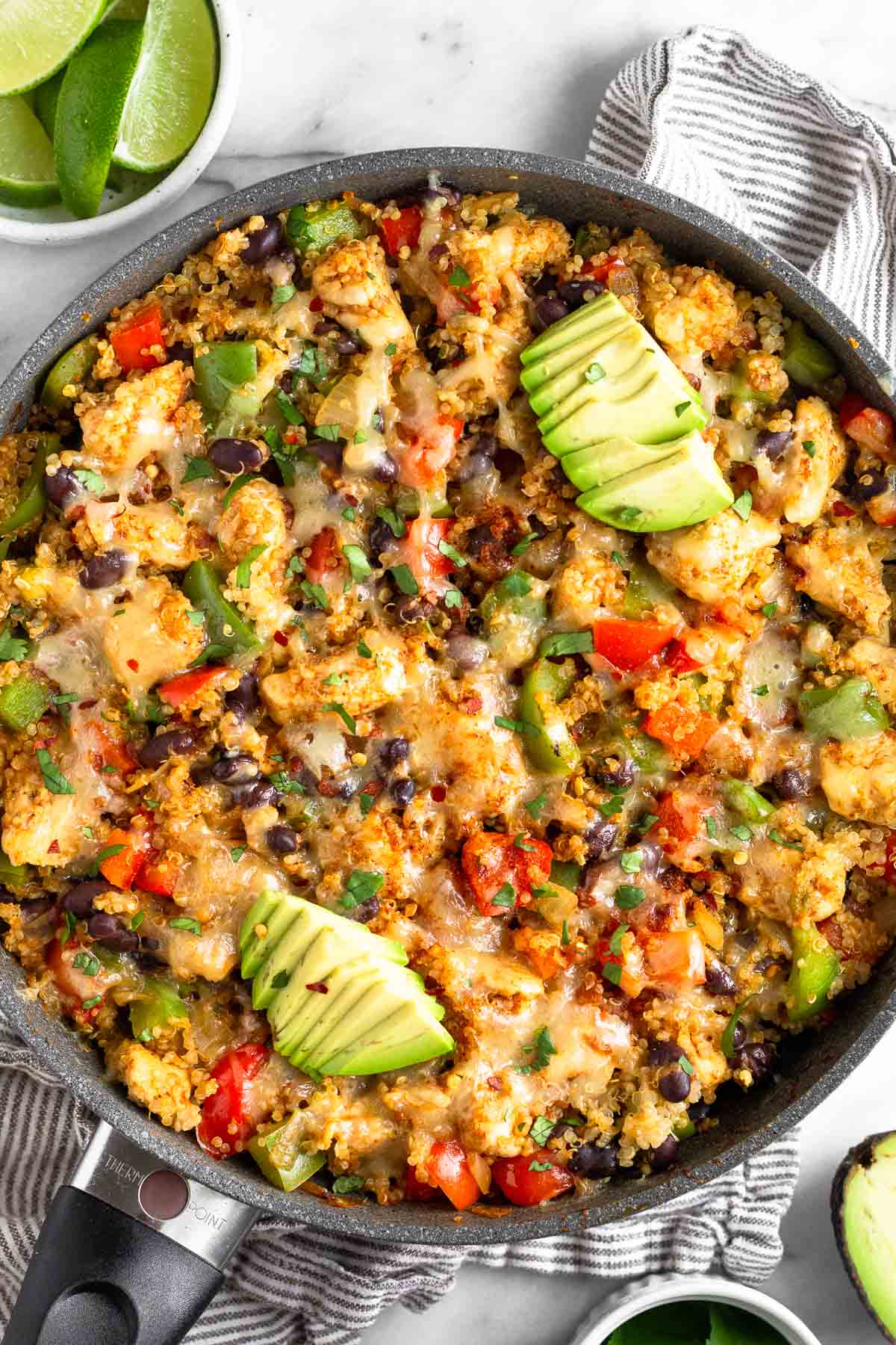 Chicken quinoa skillet in a large pan with veggies and topped with melted cheese topped with avocado. Around the pan is an half of avocado and a bowl of sliced lime.