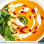 Bowl of curried butternut squash soup topped with coconut milk, hot sauce, sliced jalapenos, cashews, and cilantro. Behind it is another bowl and a clove of garlic and fresh ginger.