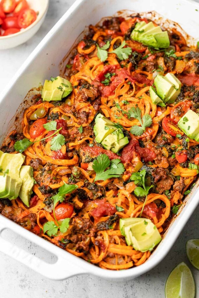 Beef taco casserole with ground beef, veggies, and sweet potato noodles in a large baking dish. It is topped with avocado and cilantro.