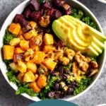 Vegan power bowl filled with butternut squash, parsnips, beets, kale, avocado, walnuts, and dried cranberries and topped with a tahini dressing. Next to is is another bowl and small dish of walnuts.