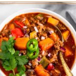 Bison chili in a bowl with a spoon in it garnished with cilantro and jalapeños.