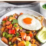Chicken hash with sweet potatoes, peppers, and bacon on a plate. It is topped with a fried egg and avocado slices.