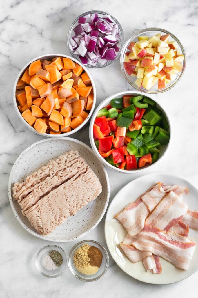 White counter with a bowl of diced red onion, a bowl of diced apples, a bowl of diced peppers, a plate of raw bacon, two bowls of spices, a plate of raw ground chicken, and a bowl of diced sweet potatoes.