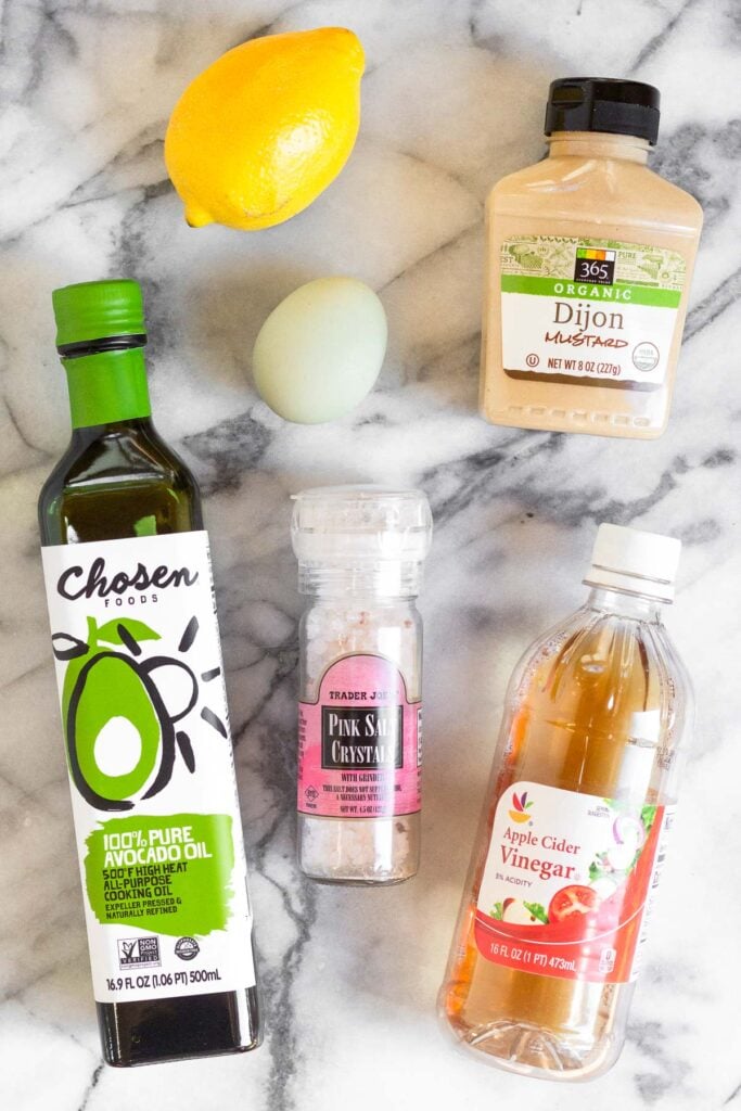 Overhead shot of a lemon, bottle of mustard, bottle of apple cider vinegar, bottle of salt, bottle of avocado oil, and an egg on white counter top.