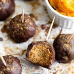 Pumpkin protein cake pops on a baking sheet with a small ramekin of peanut on it. They have toothpicks in them and one of them has a bite taken out of it.