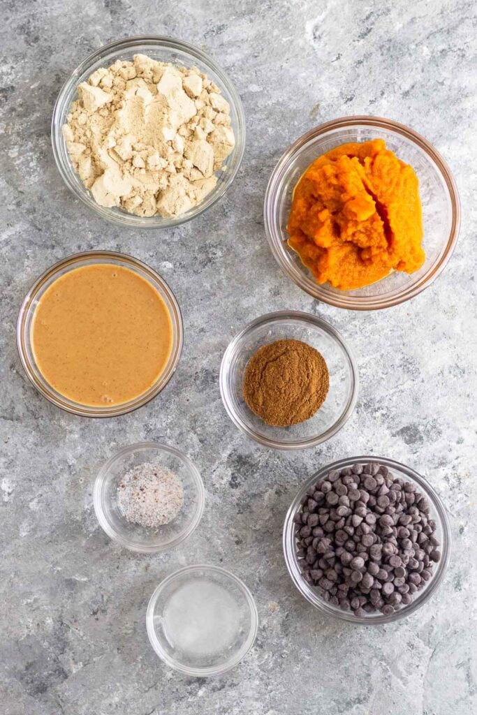 Overhead shot of a bowl of protein powder, bowl of pumpkin puree, bowl of spices, bowl of chocolate chips, bowl of coconut oil, bowl of salt, and a bowl of peanut butter.
