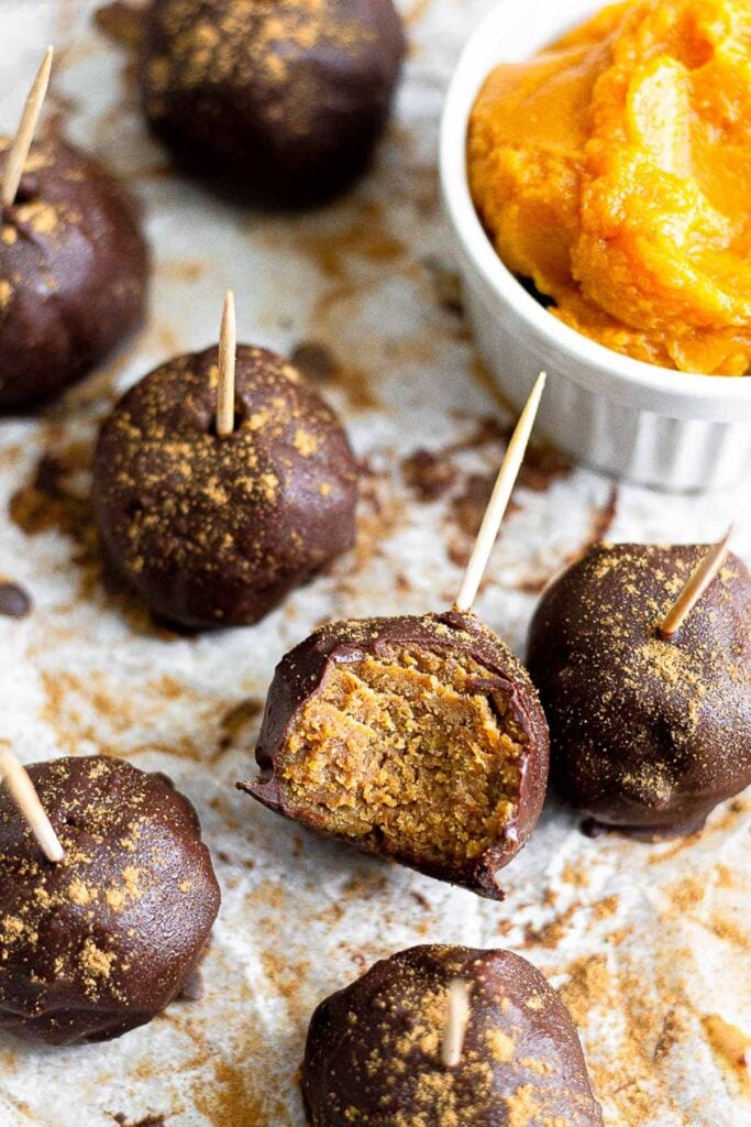 Pumpkin protein cake pops on a baking sheet with a small ramekin of peanut on it. They have toothpicks in them and one of them has a bite taken out of it.