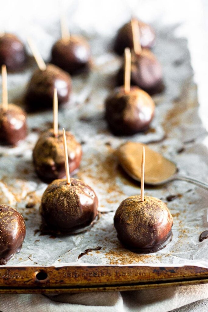 Pumpkin cake pops on a baking sheet sprinkled with pumpkin spice and tooth picks in them. There is also a spoon full of peanut butter on the sheet.