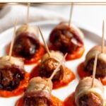 Small white plate with dollops of red pepper sauce on it. On top of the sauce are chorizo-stuffed bacon-wrapped dates. In the background is a sheet pan of more bacon wrapped dates.