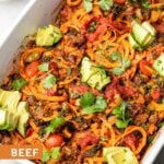 Close up of a large pan filled with ground beef taco casserole topped with cilantro and avocado. Behind it is a small bowl of diced tomatoes.