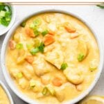 Bowl filled with buffalo cauliflower soup. it is garnished with green onions. Around it is another bowl of soup, bowl of green onions, and sliced lemons.