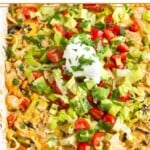 Chicken taco casserole in a large baking dish topped with tortilla chips, shredded lettuce, diced tomatoes, diced avocado, sour cream, and cilantro.