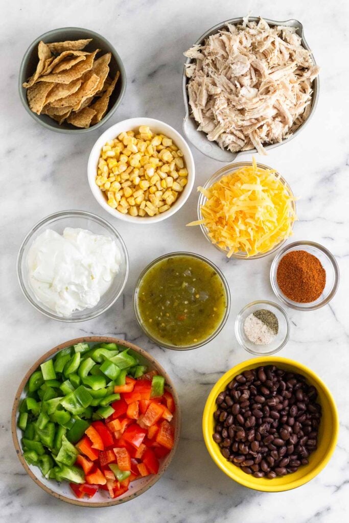 A bowl of shredded chicken, a bowl of shredded orange cheese, a small bowl of taco seasoning, a small bowl of salt and pepper, a bowl of black beans, a bowl of diced red and green bell peppers, a bowl of salsa verde, a bowl of greek yogurt, a bowl of frozen corn, and a bowl of tortilla chips on a marble counter.