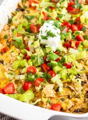 A close up of chicken taco casserole in a large baking dish topped with tortilla chips, shredded lettuce, diced tomatoes, diced avocado, sour cream, and cilantro.
