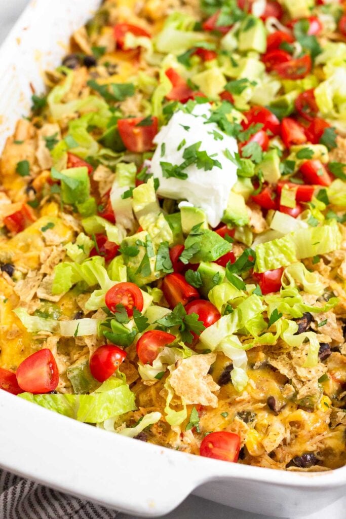 A close up of chicken taco casserole in a large baking dish topped with tortilla chips, shredded lettuce, diced tomatoes, diced avocado, sour cream, and cilantro.