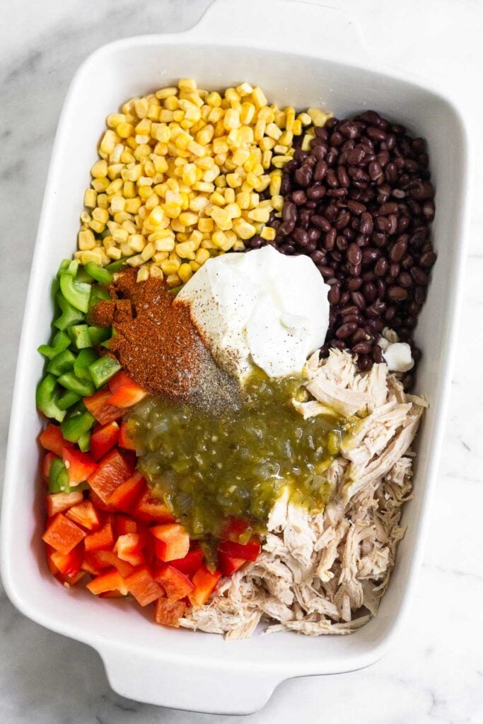 Large baking dish filled with individual piles of frozen corn, black beans, shredded chicken, diced bell peppers, salsa verde, greek yogurt, and spices before it is mixed together.