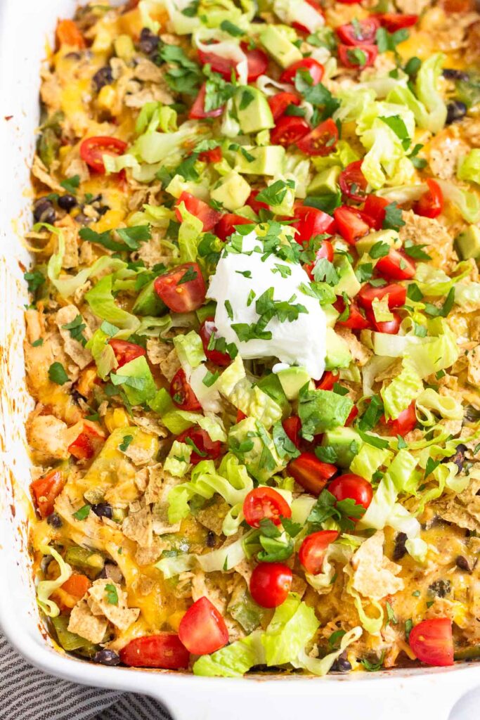 Taco chicken casserole in a large baking dish topped with tortilla chips, shredded lettuce, diced tomatoes, diced avocado, sour cream, and cilantro.