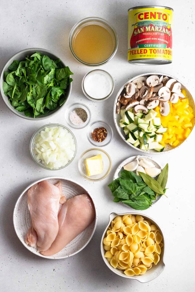 White counter with a jar of chicken broth, a can of canned tomatoes, a bowl of chopped veggies, a plate of fresh herbs and garlic, a bowl of uncooked pasta, a plate of raw chicken breasts, a bowl of diced onion, a bowl of butter, two bowls of spices, a jar of coconut milk, and a bowl of spinach.