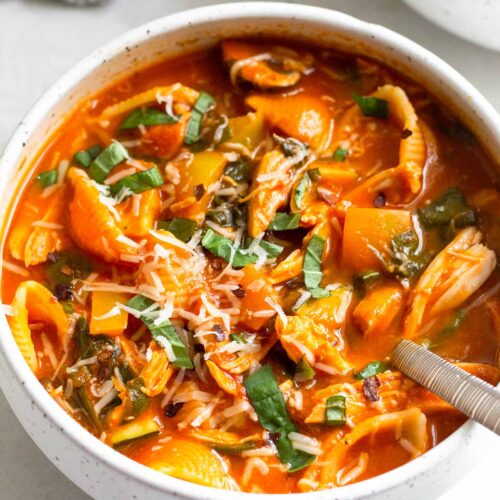 A bowl of chicken tomato soup topped with fresh herbs and cheese. A spoon is coming out of the bowl with a kitchen towel and another bowl behind it.
