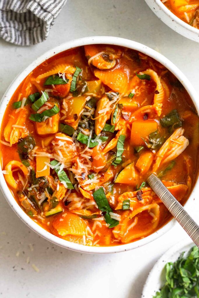 A bowl of chicken and tomato soup topped with herbs and parmesan cheese with a spoon in it. Next to it is a plate of fresh herbs, another bowl of soup, and a kitchen towel.