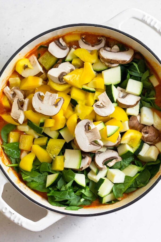 A pot of tomato soup with raw diced peppers, sliced mushrooms, and chopped spinach on top before it is mixed in.