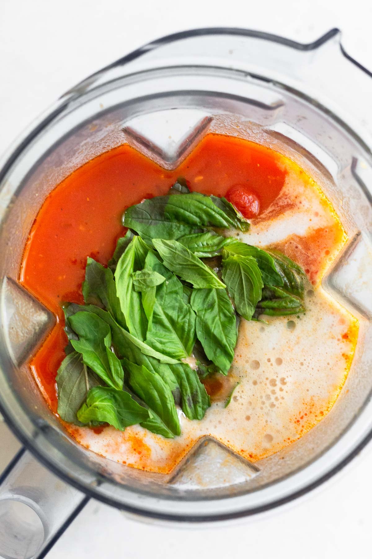 A blender filled with canned tomatoes, broth, coconut milk, and fresh basil.