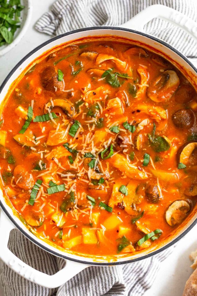 Tomato chicken soup in a large pot topped with fresh herbs and parmesan cheese. It is sitting on a kitchen towel with a plate of herbs next to it.