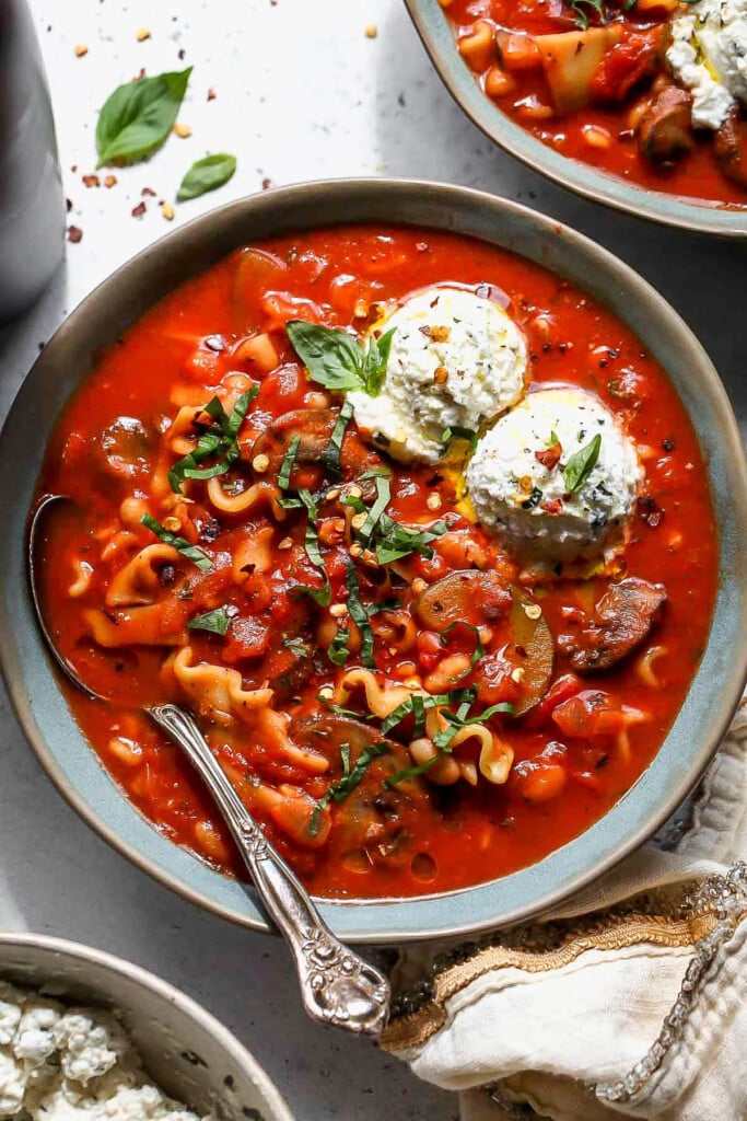 High protein vegetarian lasagna soup in a bowl topped with ricotta and fresh herbs. A spoon is in the bowl as well. Around the bowl is another bowl of soup and a linen towel.