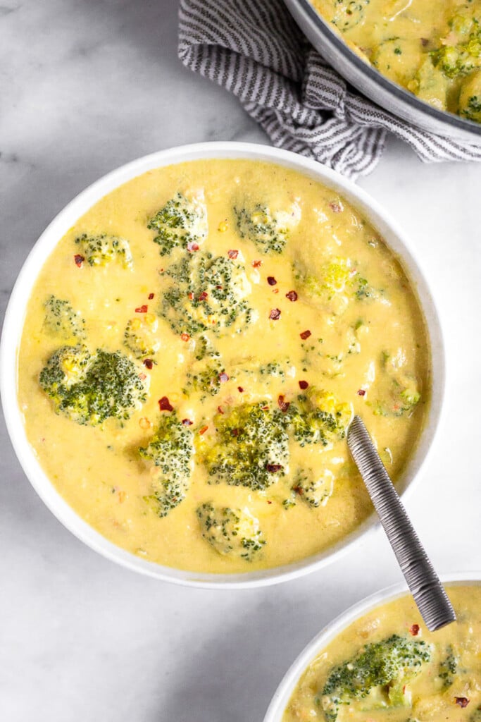 A bowl filled with vegan broccoli cheese soup topped with red pepper flakes with a spoon in it. Next to it is another bowl of soup and a pot of soup.