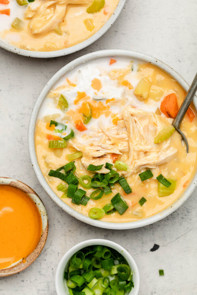 A bowl of creamy buffalo chicken soup topped with green onions. Around it is another bowl, a bowl of hot sauce, and a bowl of green onions.