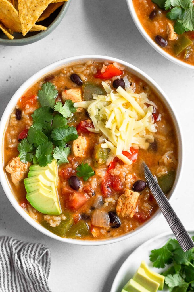 A bowl of healthy high protein soup with chicken, veggies, and beans. It is topped with avocado, shredded cheese, and cilantro and a spoon is in the bowl. Around the bowl is a plate of avocado and cilantro, another bowl of soup, a bowl of tortilla chips, and a striped towel.