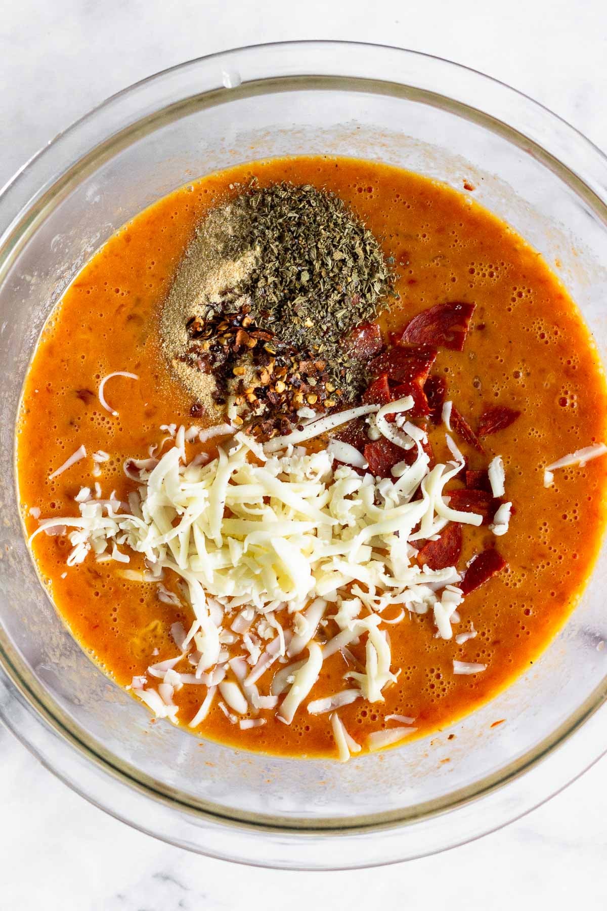 A bowl of whisked eggs with pizza sauce, shredded cheese, spices, and sliced pepperoni.