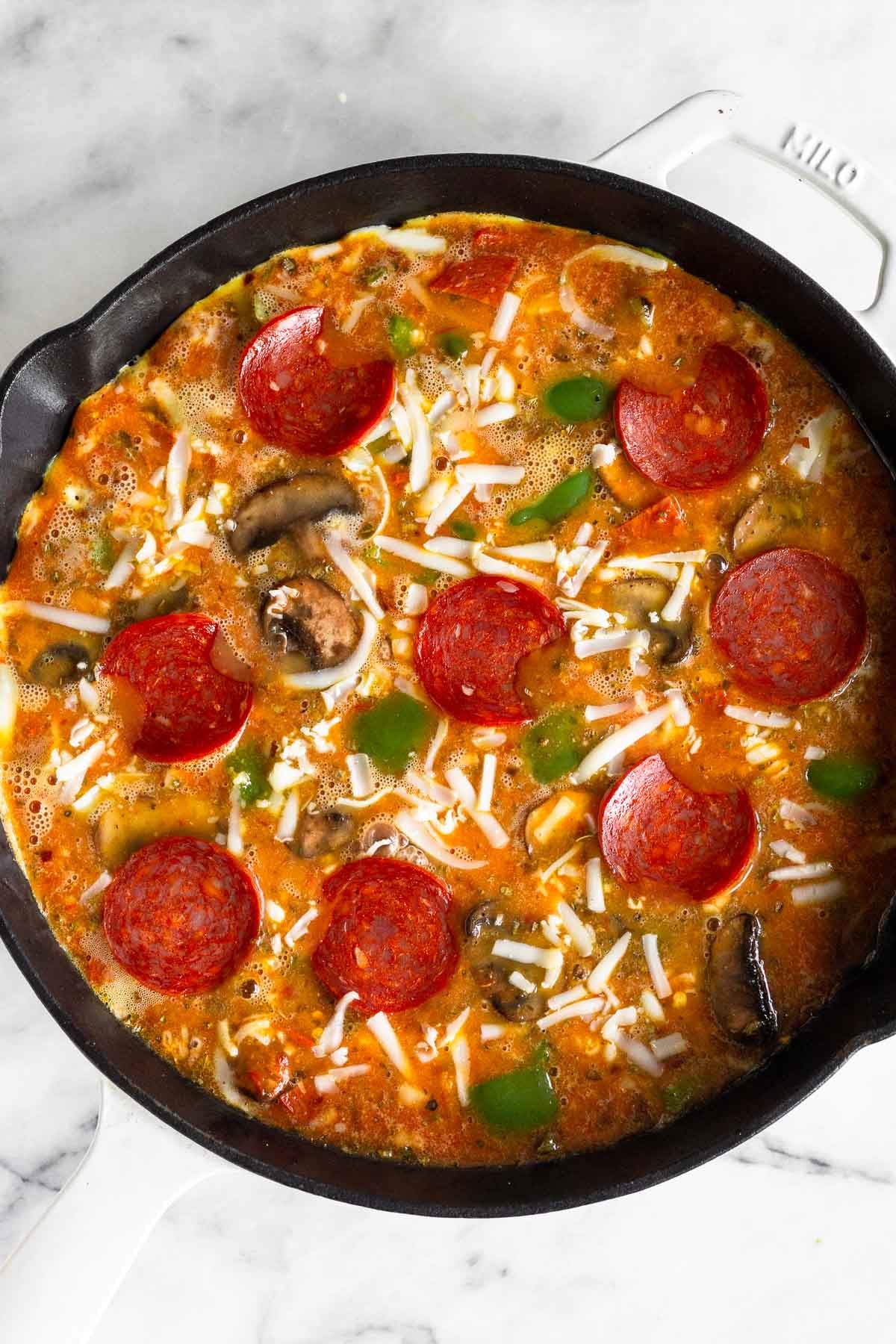 White cast iron pan filled with a mixture of eggs, cheese, veggies, and pepperoni before it is cooked.