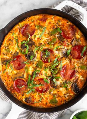 Pepperoni pizza frittata topped with fresh basil in a large white cast iron skillet. It is on a kitchen towel with a ramekin of basil next to it.