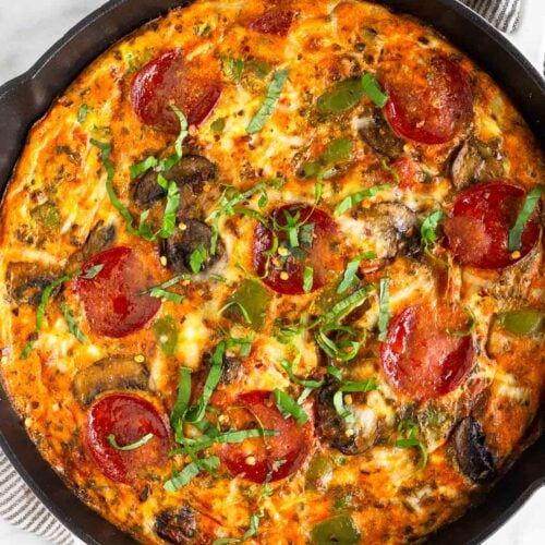 Pepperoni pizza frittata topped with fresh basil in a large white cast iron skillet. It is on a kitchen towel with a ramekin of basil next to it.